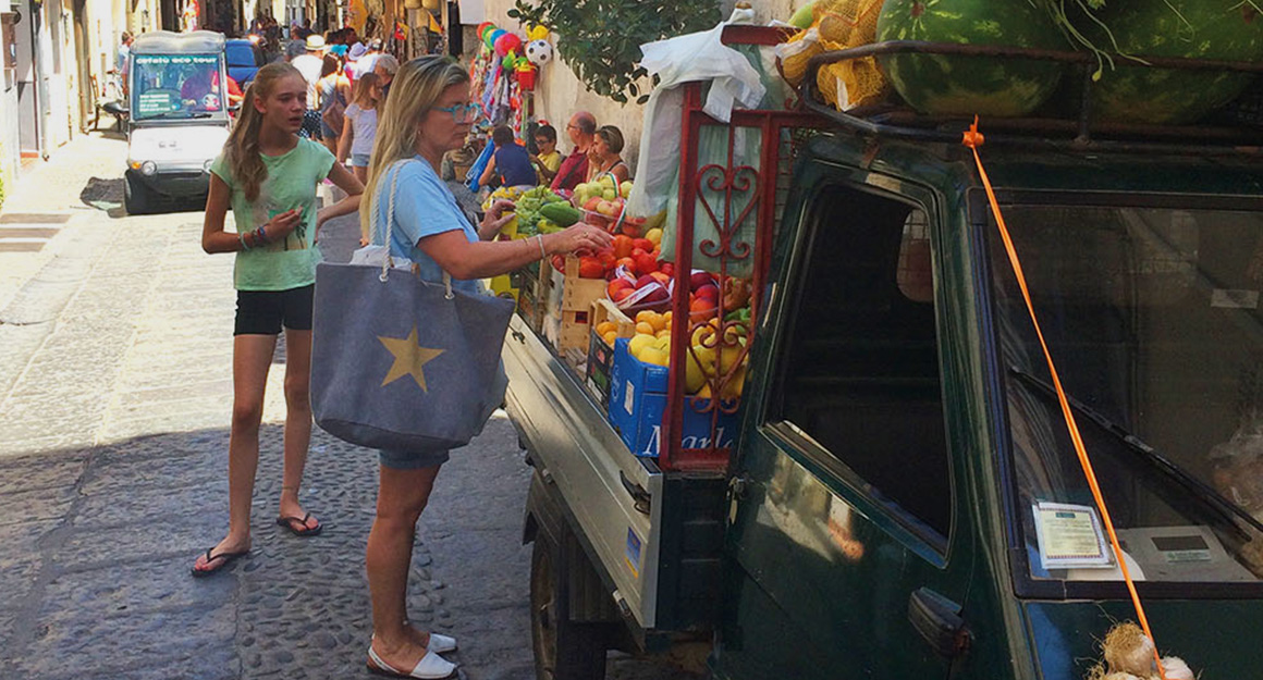 Walking through Cefalù in Sicily: lots of people in a narrow, sunny street with small vans.