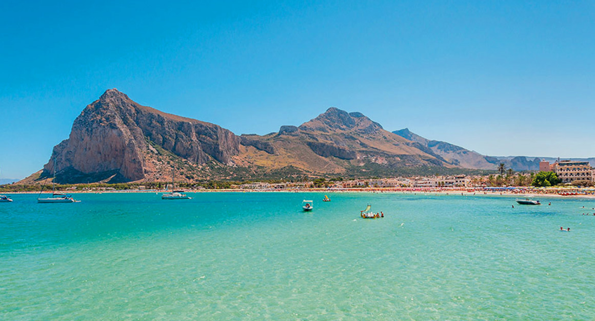 A light blue, spacious bay with a few boats and a large hilly island in the background.