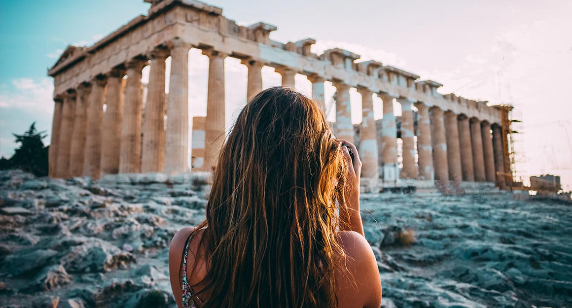 A woman photographs the ancient temple of Athens at sunset.