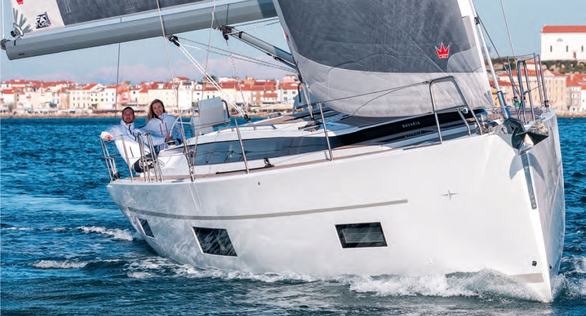 A sailing yacht from the front on the water with two people on board and a city coast.